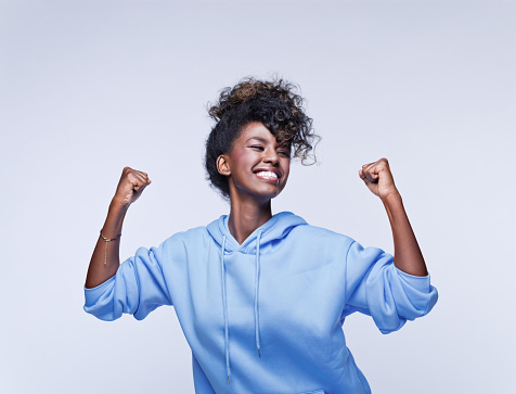 Portrait of happy african young woman wearing blue hoodie, cheering with clenched fists. Studio shot on grey background.