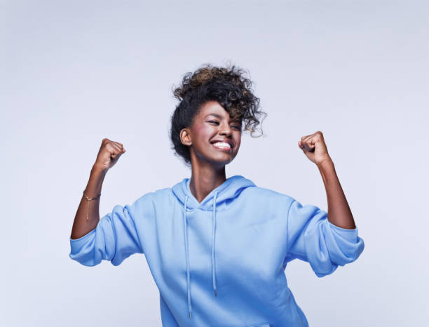 Excited young woman in blue hoodie Portrait of happy african young woman wearing blue hoodie, cheering with clenched fists. Studio shot on grey background. black woman hair bun stock pictures, royalty-free photos & images