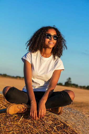 Beautiful mixed race African American female teenager young woman sitting on a hay bale in a field wearing white t-shirt and sunglasses at sunset