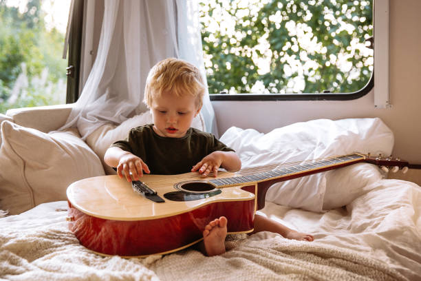 cute chłopiec grać na gitarze siedzieć na łóżku w przyczepie rv. urocze małe dziecko bawić podróży na camper - class a motorhome zdjęcia i obrazy z banku zdjęć