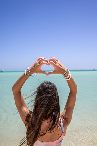 Young woman standing back turned in front of turquoise sea and making heart with hands above her head, Orange bay, Red sea, Hurghada, Egypt.