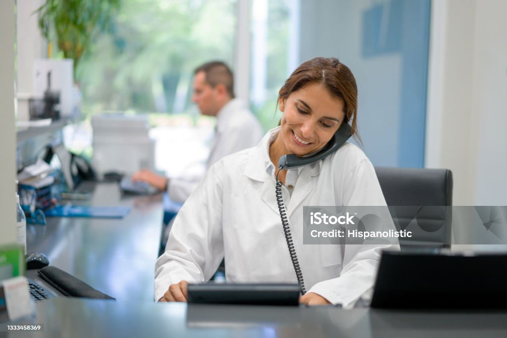 Receptionist working at a doctor's office and talking on the phone Latin American woman working as a receptionist at a doctor's office and talking on the phone -healthcare and medicine services concepts Using Phone Stock Photo