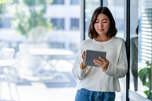 Young business woman using a digital tablet. She is casually dressed and smiling with a window behind her.