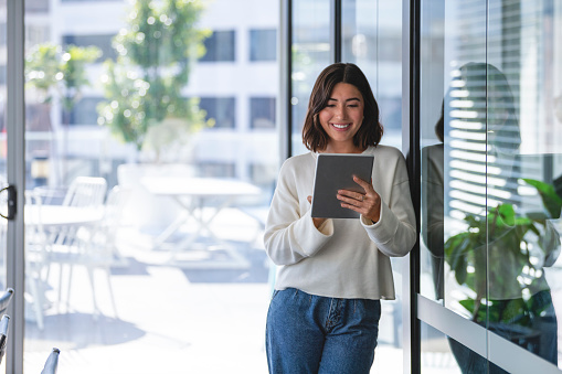 Young business woman using a digital tablet. She is casually dressed and smiling with a window behind her.