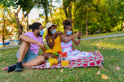A Happy Family of African-American Ethnicity With Protective Face Masks is on a Picnic in a Nature. They are Enjoying in Delicious Tropical Fruit and Having a Lot of Fun While Taking a Selfie Photos During a Corona Virus Time.