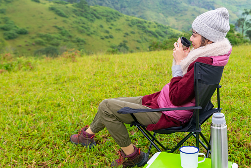 Asian woman sitting on outdoor chair with drinking hot coffee and looking to beautiful nature of mountain. Beautiful female relax and enjoy outdoor lifestyle hiking and camping in summer vacation.