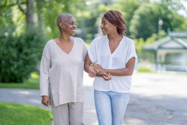 Bonding with mother who has cancer An older woman with hair loss walks down a sunny sidewalk with her daughter who is holding her arm lovingly. black mother and adult daughter stock pictures, royalty-free photos & images