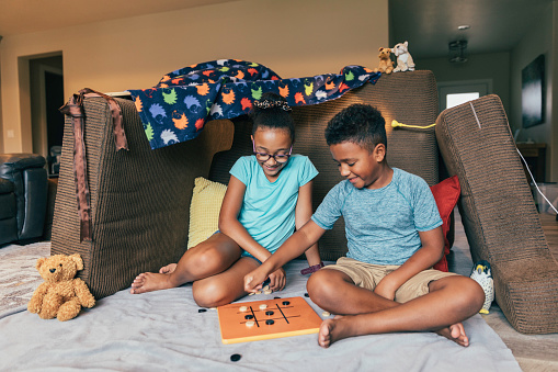A young brother and sister are playing a fun game of tic-tac-toe outside of their couch fort, constructed out of couch cushions. They love spending time together as family.