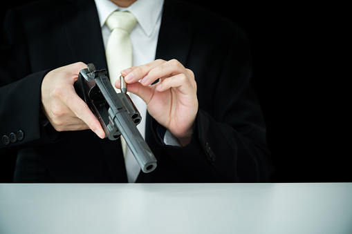 Unrecognizable Asian businessman draw the gun from gun holder inside his suit close up. Businessman or agent in black suit showing the revolver handgun on the table.