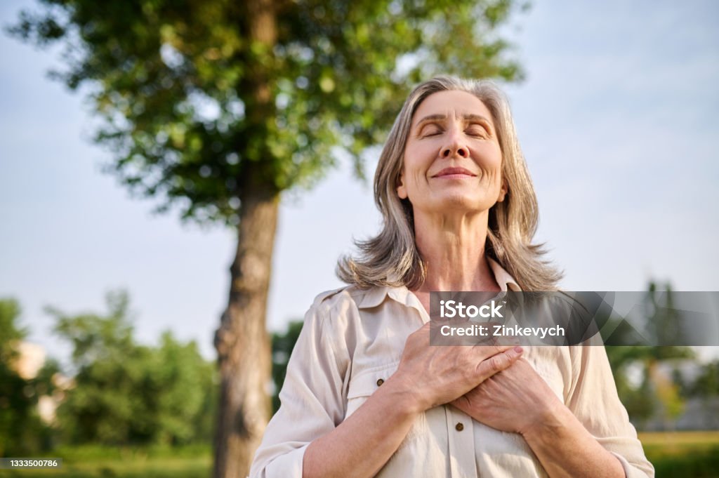 Happy woman folded hands on her chest Calmness. Happy adult smiling woman with closed eyes folded hands on her chest in summer park Healthy Lifestyle Stock Photo