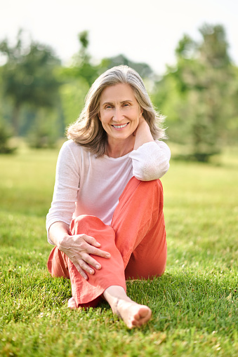 Good mood. Smiling adult barefoot woman with gray hair in casual clothes sitting on grass on summer day