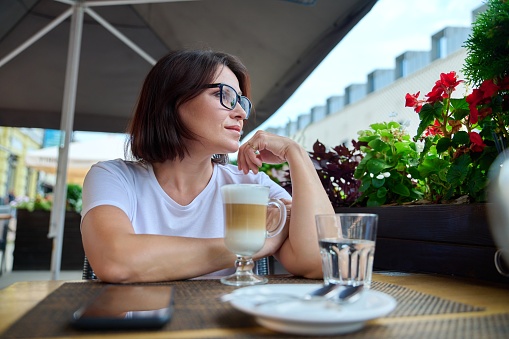 Portrait of a smiling middle-aged woman looking to the side in profile, 40s female wearing glasses sitting in an outdoor cafe with cup of coffee. Lifestyle, urban style, middle aged people concept