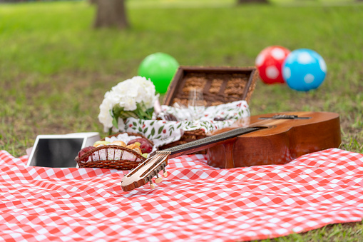 Red and white checkered picnic tablecloth lying on the lawn with a guitar, a basket with food, a tablet and some colored balls.