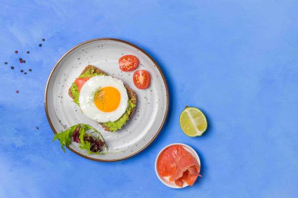 Toast with guacamole, fried eggs and slices of smoked salmon, on a gray plate with lettuce and tomatoes. On a blue background, horizontal with space stock photo