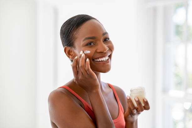 Happy black mature woman applying cream on face Middle aged woman applying hydrating moisturizer on her face, daily routine skin care. Smiling middle aged african woman holding little jar of skin cream and applying lotion on cheek. Beautiful young black lady applying anti-aging cream on face. face cream stock pictures, royalty-free photos & images