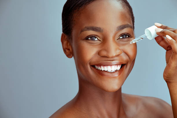 Black beauty woman dropping collagen serum moisturizer on face Portrait of african american woman applying liquid cosmetic oil for facial treatment against blue background with copy space. Beauty black middle aged woman with natural makeup holding glass dropper. Close up face of mature lady applying serum around dark circle eyes to combat wrinkles. face serum stock pictures, royalty-free photos & images
