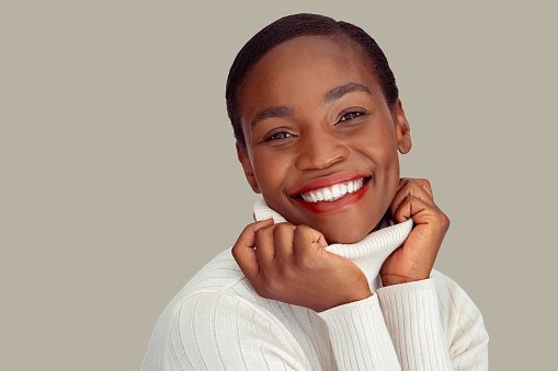 Portrait of fashionable african woman holding collar of winter clothing and smiling against white background. Cheerful mid adult black lady wearing cozy sweater against wall with copy space. Close up face of laughing and carefree woman looking at camera during christmas.