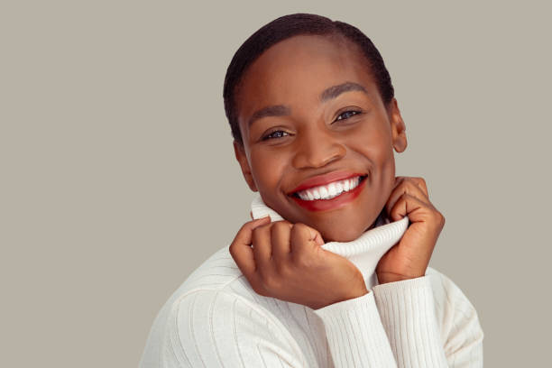 Beautiful mature woman in winter clothing laughing Portrait of fashionable african woman holding collar of winter clothing and smiling against white background. Cheerful mid adult black lady wearing cozy sweater against wall with copy space. Close up face of laughing and carefree woman looking at camera during christmas. beautiful older black woman stock pictures, royalty-free photos & images