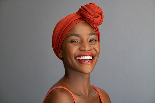 Portrait of cheerful african mature woman with headscarf against grey wall. Portrait of middle aged black woman laughing with copy space. Happy smiling black lady wearing traditional african scarf and looking at camera.