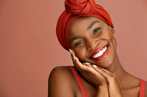 Portrait of beauty and lovely woman with ethnic headscarf looking at camera. Confident mid adult black woman smiling while wearing red headband against red wall. Beautiful african american mid woman with flawless skin