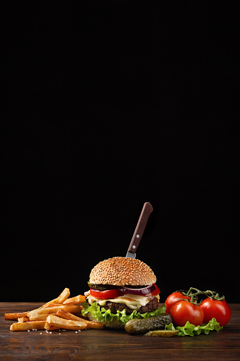 Homemade hamburger close-up with beef, tomato, lettuce, cheese, onion and french fries on wooden table. In the burger stuck a knife. Fastfood on dark background with place for your text.