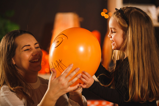 Candid shot of cheerful little girl showing her mother a drawing of a spooky Jack o' lantern face that she drew on an orange balloon as a decoration for Halloween.