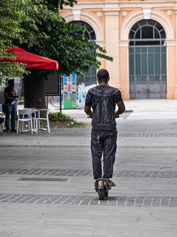 Parma, Italy - june 2021: Black Man Dressed in Gray Crossing a Public Square on the Electric Mono Scooter.