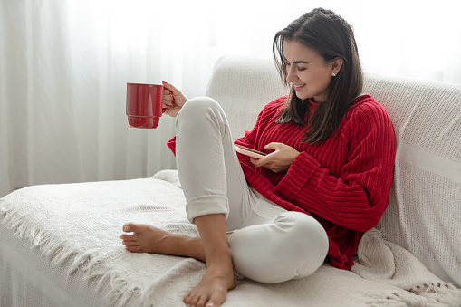 A young woman in a red sweater with a red cup in her hands looks at the phone while sitting on the couch.