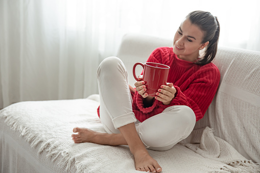 A young woman with a red cup of hot drink in a cozy red sweater is resting on the couch at home copy space.