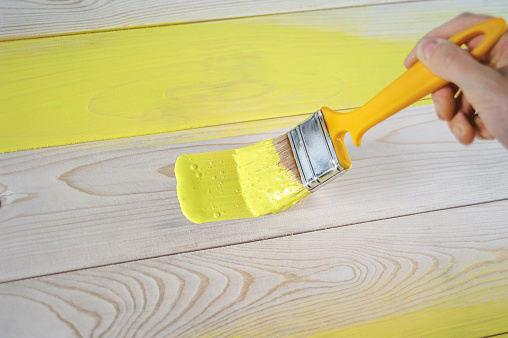 Closeup female hand with paintbrush painting a natural wooden door with yellow paint. The concept of colored bright creative interior design for a young family. How to paint a wooden surface.