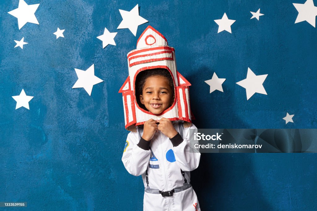 Afro american little boy in space suit playing astronaut on blue backdrop with stars. Childhood, creative, imagination Rocket afro-american little boy in space suit playing astronaut on blue background with white hand-made stars. Happy mixed race child with handcrafted spacecraft. Childhood, creative and imagination. Child Stock Photo