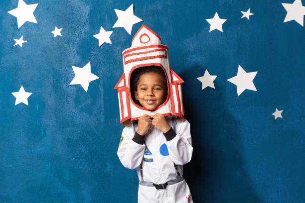 Afro american little boy in space suit playing astronaut on blue backdrop with stars. Childhood, creative, imagination Rocket afro-american little boy in space suit playing astronaut on blue background with white hand-made stars. Happy mixed race child with handcrafted spacecraft. Childhood, creative and imagination. playful stock pictures, royalty-free photos & images