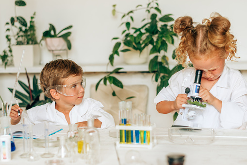 Boy and girl in white uniforms and protective glasses do chemical experiments in a home laboratory.Back to school concept.Young scientists.Natural sciences.Preschool and school education of children.