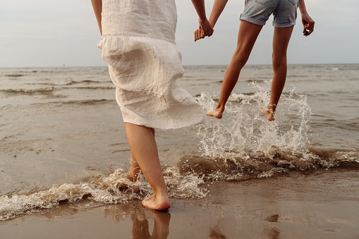 mother in white dress and daughter in shorts legs close up jumping in the sea with splashes holding hands. High quality photo