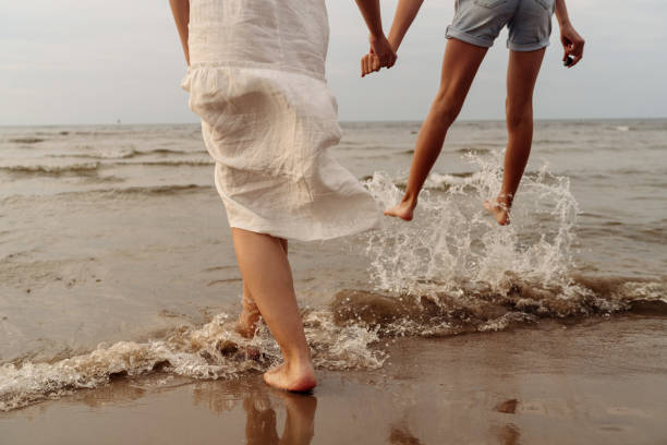 mother in white dress and daughter in shorts legs close up jumping in the sea with splashes holding hands mother in white dress and daughter in shorts legs close up jumping in the sea with splashes holding hands. High quality photo wave jumping stock pictures, royalty-free photos & images