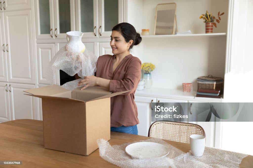 Happy Indian woman unpack box with tableware order Happy young Indian female renter or tenant settle in new home unbox parcel shopping tableware online from home. Smiling ethnic woman unpack box buying kitchenware on internet. Delivery concept. Moving House Stock Photo