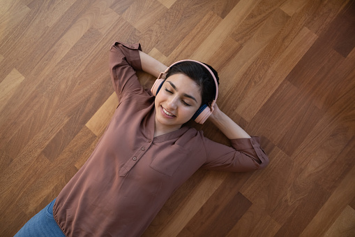 Top view of happy young Indian woman wear headphones lying on floor in living room listen to music. Smiling ethnic female in earphones have fun relax at home enjoy good quality sound on gadget.