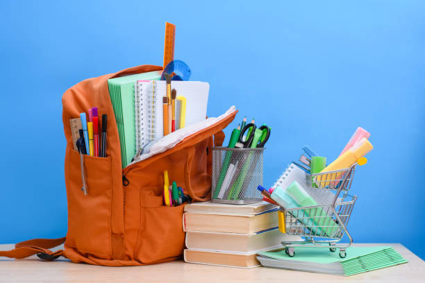 mochila de escola laranja cheia de material escolar e uma cesta de supermercado com material de escritório em um fundo azul. - material escolar - fotografias e filmes do acervo