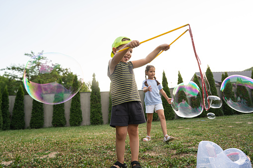 Playful and adventurous siblings, playing together and making a big soap bubble with a bubble wand, during family gathering at the backyard of country house