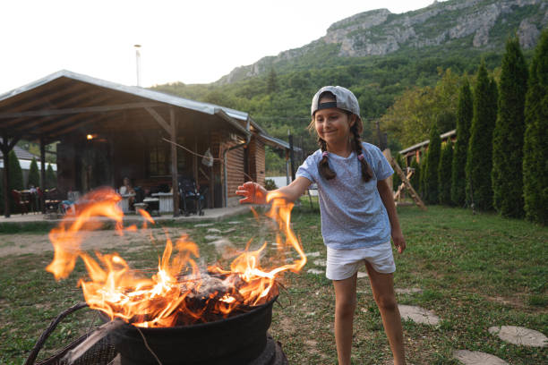 любопытная и улы�бающаяся 5-летняя девочка, бросающая ветки в пламя барбекю во время семейного сбора - 6 7 years lifestyles nature horizontal стоковые фото и изображения