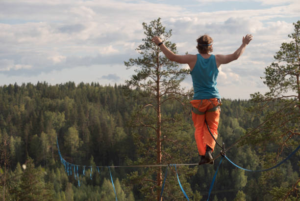 A tightrope walker walks along a long highline above the forest. Back view A tightrope walker walks along a long highline above the forest. Back view highlining stock pictures, royalty-free photos & images