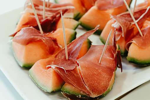 several pieces of melon with Prosciutto and wooden toothpicks, arranged on a white plate with selective focus on the foreground