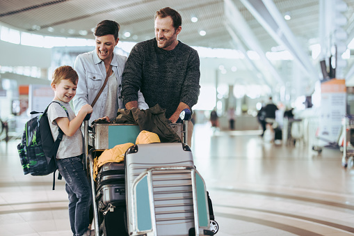 Husband and wife looking at child standing on luggage trolley at airport. Couple looking at their son at airport with luggage trolley.
