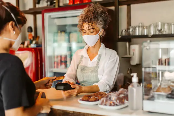 Business owner in a face mask taking a card payment from a customer. Mature businesswoman serving a customer over the counter in her cafe. Customer service during the COVID-19 pandemic.