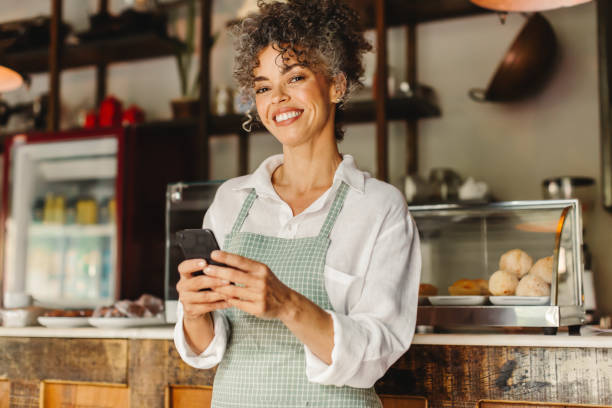 Small business owner holding a smartphone in her cafe Small business owner using a smartphone in her cafe. Mature businesswoman smiling at the camera while standing in front of the counter. Successful entrepreneur communicating with her customers online. entrepreneur stock pictures, royalty-free photos & images