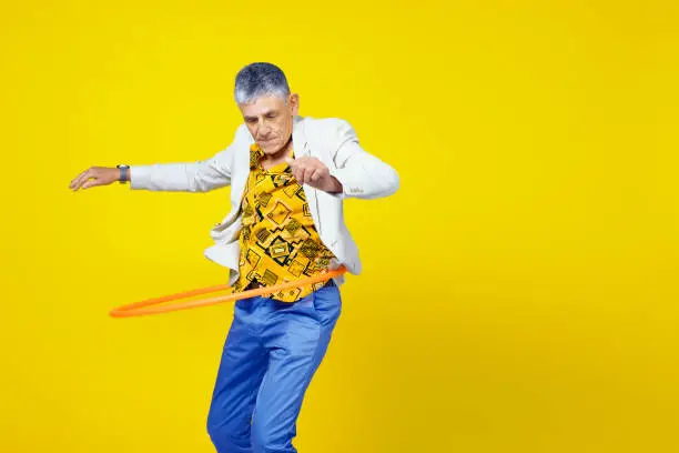 Funny and extravagant senior man in his sixties having fun and hula-hooping on a colored background