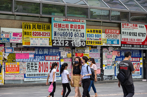 Hong Kong - August 11, 2021 : People wearing protective face masks walk past the shuttered retail space covered with property advertisements in Tsim Sha Tsui, Kowloon, Hong Kong.