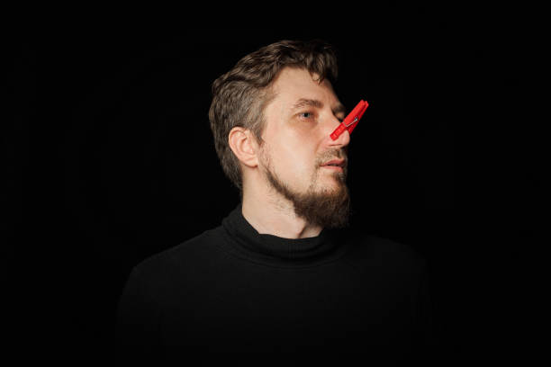 Bearded man with clothespin on nose. Bad stink, funny prankster, joke concept. Laugh at yourself. Red clothespin - symbol of self-mockery and joking character. Black background.
