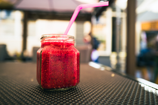 Close up of a healthy red berry smoothie, loaded with vitamins, in a glass jar with pink plastic straw on a restaurant table outdoors.