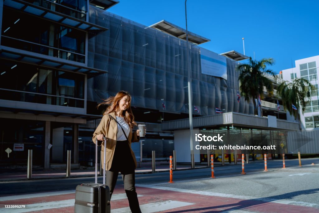 Business woman is walking inside the terminal. A young woman with coffee and a suitcase is walking on an outdoor road on the grounds of the airport. Asian and Indian Ethnicities Stock Photo